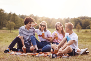 Photo of classmates have rest outdoor, sit on plaid together, being pleased to speak with each other, hold bottles of cold drink, rejoice summer vacation. People, youth, lifestyle and friendship