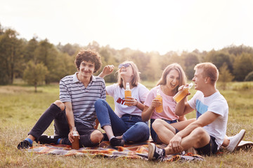 Shot of happy teenagers laugh joyfully while have picnic outdoor, drink cold energy drinks, sit crossed legs, have fun, breath fresh air, pose outside, spend weekend on nature. Young and leisure