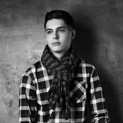 Portrait of young trendy handsome man with short dark hair wearing checkered shirt and brown trousers standing and posing against gray concrete wall