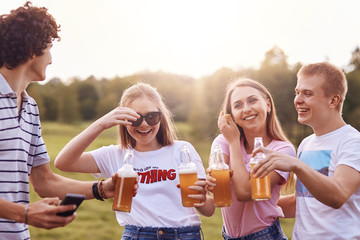Naklejka premium Summer time and leisure concept. Happy girls laugh at boy`s funny jokes, drink cold beverages, have cheerful expressions, spend spare time on summer field. Companionship and relationship concept