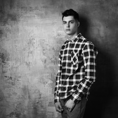 Obraz premium Portrait of young trendy handsome man with short dark hair wearing checkered shirt and brown trousers standing and posing against gray concrete wall