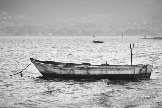 Old empty boat on shore / Black and white view of a rustic fishing boat moored on seashore on a cloudy and stormy day. It represents loneliness.