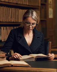 Beautiful blonde woman wearing elegant black tweed jacket and glasses sitting at desk beside bookshelf and reading book. Young gorgeous female student studying at library. Smart is new sexy concept.