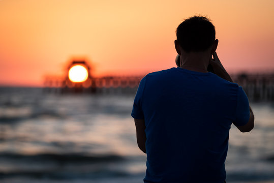 Naples, Florida pink, red and orange sunset in gulf of Mexico with sun setting inside Pier bokeh, back of young man photographer videographer taking pictures filming landscape