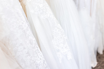 Obraz na płótnie Canvas Many wedding dresses on rack in boutique discount store, white garments hanging hangers row closeup with white lace, tulle, design