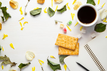 An open blank notebook and a black pencil, cup of coffee and cookies on a white wooden background among dry leaves and flower.