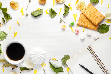 An open blank notebook and a black pencil, cup of coffee and cookies on a white wooden background among dry leaves and flower.