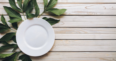 Empty white plate on light gray rustic wooden background with green leaves of peony. Top view, copy space.