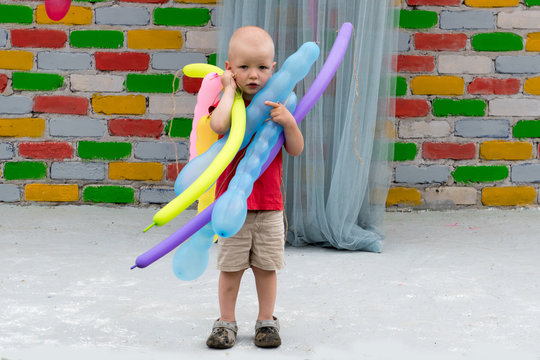 Cute boy with shaved head in red T-shirt with colored balloons against multi colored brick wall.