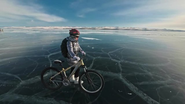 Woman is walking beside bicycle on the ice. The girl is dressed in a silvery down jacket, backpack and helmet. Ice of the frozen Lake Baikal. The tires on the bicycle are covered with special spikes