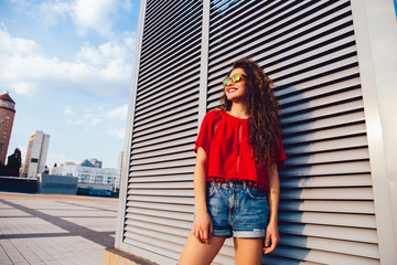 Attractive young woman in sunglasses dressed in bright blouse and shorts posing near a modern wall outdoors.