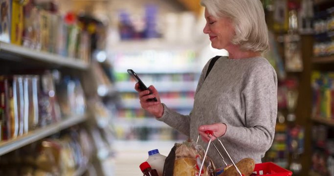 Happy Senior woman looking at grocery store aisle with cellphone in hand, Smiling older female using mobile phone app to shop for food, Retired person using diet app on smartphone in market, 4k 