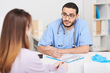 Fototapeta premium Portrait of young Middle-Eastern doctor wearing glasses talking to female patient sitting at desk in office and filling out patients card
