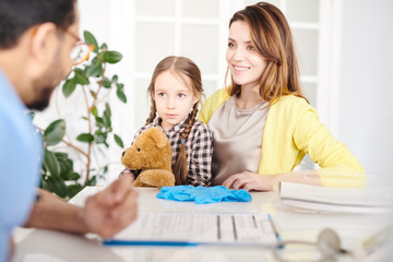 Portrait of beautiful young mother and cute little girl sitting at desk in doctors office and listening to pediatrician during visit