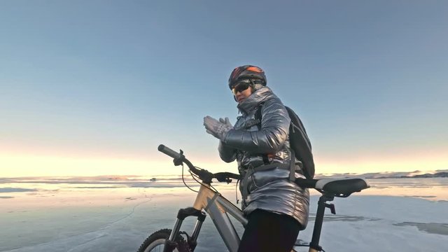 Woman is stay in bicycle on the ice. The girl is dressed in a silvery down jacket, cycling backpack and helmet. Woman is heating her hands with friction. Ice of the frozen Lake Baikal. The traveler is