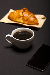 Croissant, smartphone and cup of coffee on black background