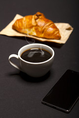 Croissant, smartphone and cup of coffee on black background