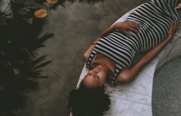 Young African-American pregnant woman in a striped dress and with curly Afro hair is resting in the summer dusk on the marble bench next to the pond with waterlilies and reflections of the palm trees