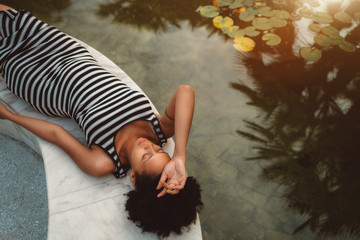 Tired beautiful pregnant African-American girl with curly afro hair is resting and laying on the marble bench near the pond with waterlilies on a sunny summer morning, palms silhouettes in the water - Powered by Adobe