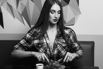 Gorgeous seductive woman dressed in unbuttoned checkered shirt sitting at table and eating delicious steak with knife and fork. Young beautiful female model at steakhouse or meat restaurant.