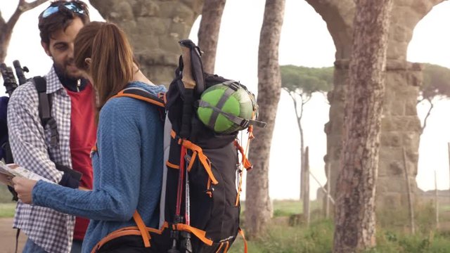 Happy young couple backpackers tourists reading map guide for directions lost near roman aqueduct arches in parco degli acquedotti park ruins in rome at sunrise with guitar and sleeping bag slow