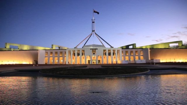 Australian national public building of parliament in Canberra on capitol hill at sunset with bright illumination and waving australian flag on flagpole.
