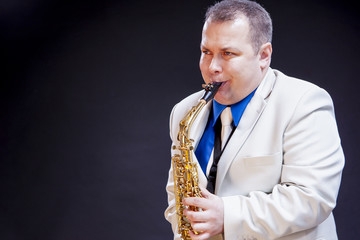 Fototapeta premium Music Concepts. Expressive Mature Caucasian Male Saxophone Player Posing in White Suite With Sax. Against Black Background.