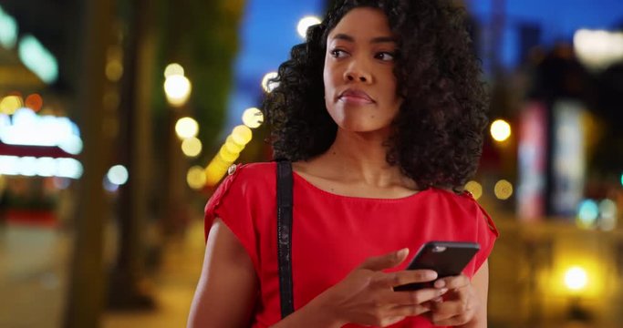 Smiling African American woman text messaging with cell phone on city street at night, Happy young black Millennial using smartphone waiting for ride on warm summer evening, 4k