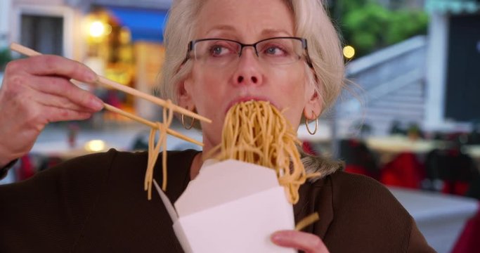 Slow motion portrait of mature woman eating Chinese takeout, An older female eating noodles with chopsticks, 4k