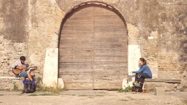 Happy young couple backpackers tourists playing guitar and singing sitting near wooden doorway of medieval building in parco degli acquedotti park ruins in rome at sunrise with map guide slow motion