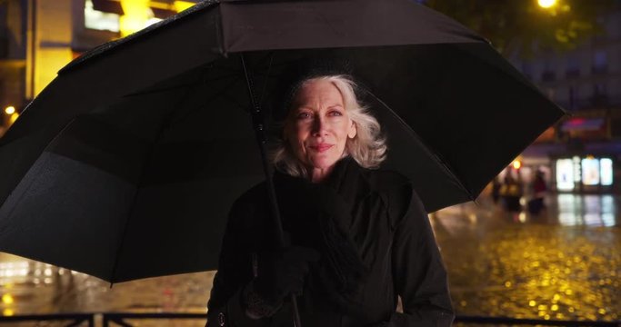 Happy senior woman in Paris smiling under umbrella on rainy night in the city, Beautiful mature Caucasian tourist waiting for ride in the rain, Retired female on vacation during winter, 4k