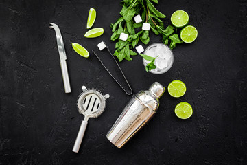 Ingredients and crockery for making mojito. Slices of lime, mint, sugar cubes, glass with ice cubes, shaker, strainer on black background top view copy space
