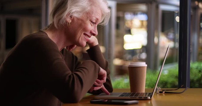 Slow motion smiling senior woman using laptop computer in restaurant or cafe at night, Happy older woman browsing internet in coffee shop with wifi, 4k