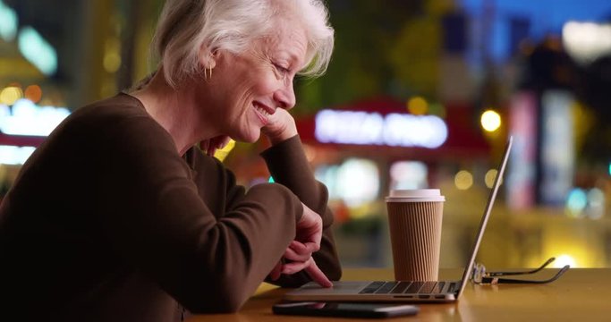 Slow motion smiling senior woman using laptop computer in restaurant or cafe at night, Happy older woman browsing internet in coffee shop with wifi, 4k