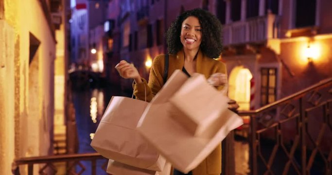Stylish young black woman tourist shopping in Venice at night stops to dance and spin playfully, Happy young fashion forward African woman with bags on vacation in Italy, 4k