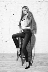 Young beautiful woman with long straight blonde hair in blue jeans and bright blouse sitting on white high bar chair with crossed legs and looking in camera.