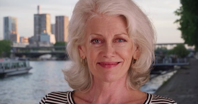 Close up portrait of attractive older white woman wearing striped shirt smiling at camera in Paris by the River Seine, Retired Caucasian woman smiles in France, 4k