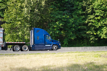 Blue big rig semi truck profile going with semi trailer on the road with green trees