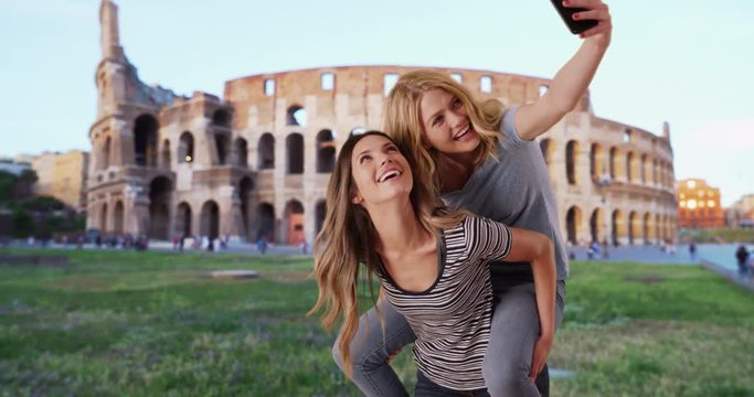 Attractive blonde Caucasian female riding piggyback on her friend's back takes a selfie near the roman Colosseum, Pretty brunette in Rome gives friend piggyback ride to take selfies, 4k
