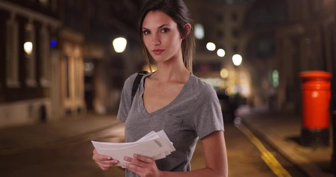 Portrait of Caucasian woman holding stack of mail in city street at night, Millennial girl with letters in hand looking at camera standing alone in urban street, 4k