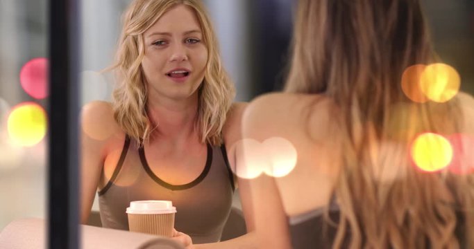 Cute blonde fitness woman talking with friend at table with yoga mat indoors coffee shop, Healthy yoga practitioner talking with friend inside building facing street with bokeh reflection, 4k