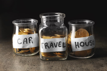 Glass jars with coins for car, house and travel on table against dark background