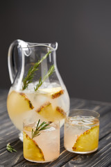 jug and two glasses of lemonade with pineapple pieces, ice cubes and rosemary on grey wooden tabletop