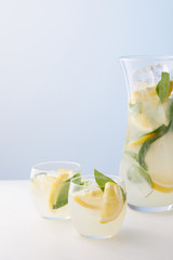 jug and two glasses of lemonade with mint leaves, ice cubes and lemon slices on blue background