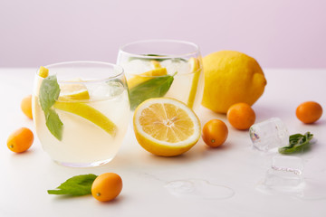 two glasses of lemonade with mint leaves, ice cubes and lemon slices surrounded by kumquats and lemons on purple background