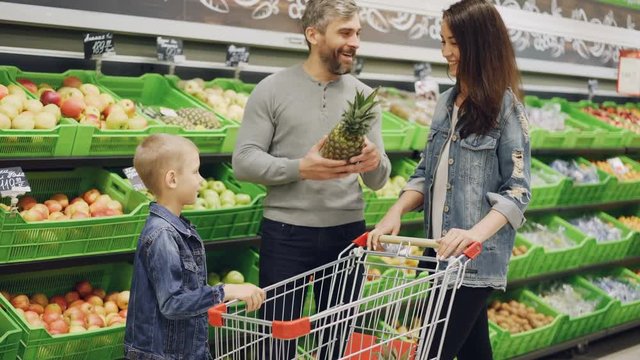 Father, mother and child are choosing pineapple in grocery store touching and smelling it showing thumbs-up. Shelves with delicious fruit and shopping trolley are visible.
