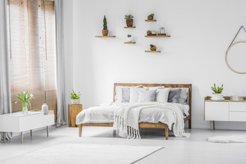Wooden shelves with plants above a comfortable double bed in a spacious apartment room interior with white furniture and walls