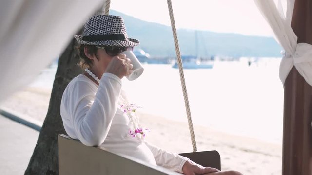 Woman in aged drinks tea on swing resting on the beautiful beach and swings. slow motion. 3840x2160
