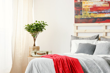 Real photo of a bed with grey and white bedding and pillows standing next to a shelf with a plant in a bedroom interior with a painting on a wall