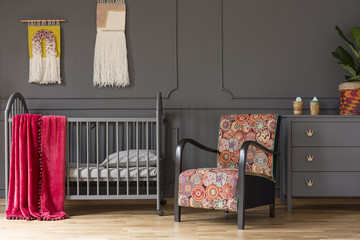 Patterned armchair and grey cabinet next to kid's bed with red blanket in bedroom interior. Real...